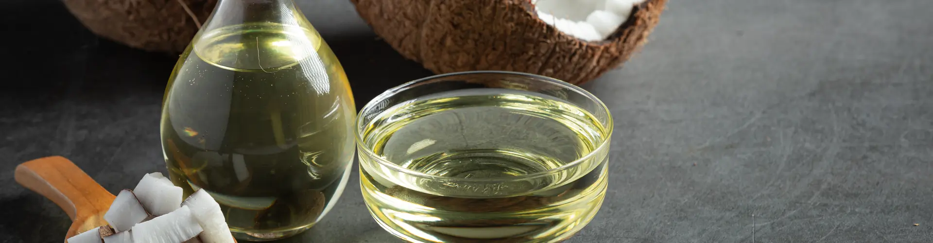 A glass bowl of coconut oil sits on a surface next to halved coconuts, a coconut piece, and a palm leaf in the background.